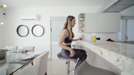Young-blonde-woman-in-sportswear-sitting-in-a-luxurious-kitchen-to-have-a-juice-for-breakfast
