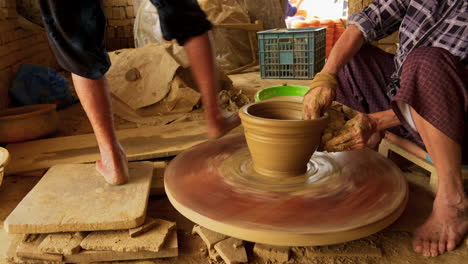 Two-Asian-women-collaborate-on-making-pottery,-turning-a-foot-powered-table-and-crafting-a-new-art-piece