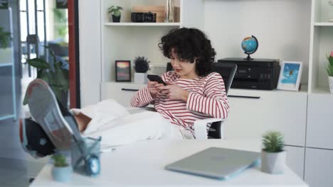 Portrait-of-a-beautiful-young-curly-haired-woman-sitting-at-a-desk,-propping-her-feet-on-the-table,-playing-an-engaging-game-on-her-phone,-and-smiling