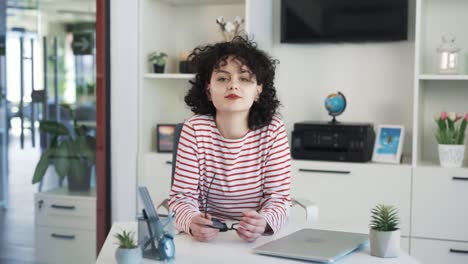 Portrait-of-a-beautiful-young-curly-haired-woman-sitting-at-a-desk-with-a-laptop-in-a-spacious-bright-office,-taking-off-her-glasses-and-smiling-at-the-camera