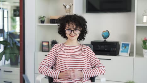 A-portrait-of-a-beautiful-young-curly-girl-sitting-at-a-table-with-a-laptop-in-a-spacious-light-office-by-handing-hair-and-glasses-for-vision-and-smiles-at-the-camera
