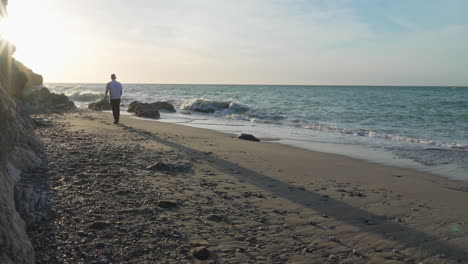 man-standing-on-beach,-walks,-picks-up-stone-during-sunny-morning,-sea-waves-wash-onshore