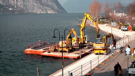 Time-lapse-construction-work-on-waterfront,-excavator-building-support-pillar-in-lake-water-surface,-heavy-industrial-equipment-on-floating-construction-platform-in-Italy,-Iseo