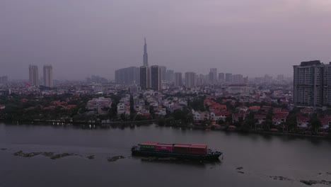 Container-boat-carrying-cargo-on-Saigon-River,-Vietnam-at-sunset-with-drone-view-of-water-and-city-skyline