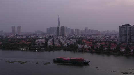 Barco-Contenedor-Que-Transporta-Carga-En-El-Río-Saigón,-Vietnam-Al-Atardecer-Con-Vista-De-Drones-Del-Agua-Y-El-Horizonte-De-La-Ciudad