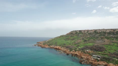 Vista-Aérea-Giratoria-Sobre-Una-Costa-Rocosa-Naranja-Con-Agua-Turquesa-En-La-Base-De-Una-Colina-Verde,-Isla-De-Gozo,-Malta