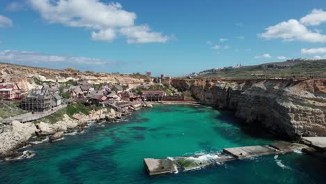 Drone-View-Over-Popeye-Village-Theme-Park-In-Malta-With-Rocky-Shoreline-And-Turquoise-Water