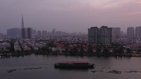 Container-boat-carrying-cargo-on-Saigon-River,-Vietnam-at-sunset-with-panning-view-of-water-and-city-skyline
