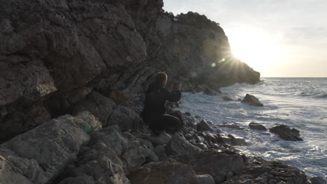 Man-squatting-taking-a-photo-of-morning-sun-over-sea,-he-walks-away-along-cliff-side