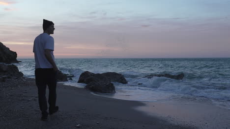 Man-in-white-shirt-walks-along-the-ocean-coast-during-nightfall,-takes-photo-of-sea