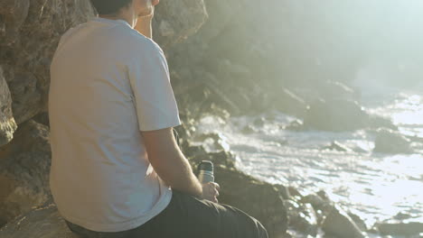 Man-drinks-thermos-coffee-looking-out-into-the-sea,-as-morning-sun-shines-on-him-and-waves-crush-in-rocks-near-by