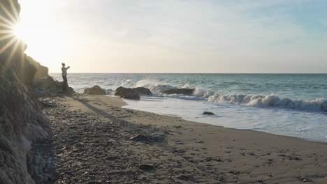 Mann-Verspielt-Weit-Am-Strand,-Wirft-Steine,-Hüpft-Herum,-Lange-Schatten-Wirft-Hinterher,-Meereswellen-Spülen-An-Land