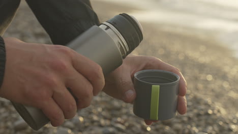 Man-opens-thermos,-pours-coffee-at-beach-with-sea-waves-golden-bay-blurry-background