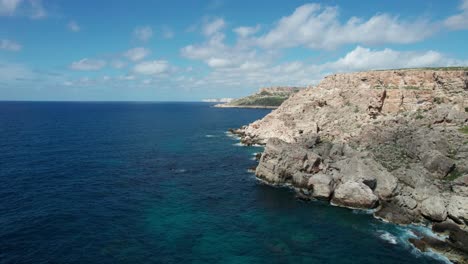 Aerial-View-Of-A-Rocky-Shoreline-In-Malta,-With-Clear-Blue-Sky-And-Thick-White-Clouds