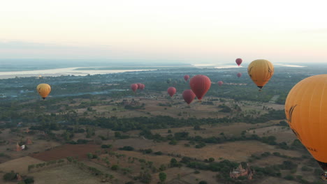 Toma-Aérea-Hacia-Atrás-Que-Muestra-Un-Grupo-De-Globos-Aerostáticos-Que-Vuelan-Sobre-El-Paisaje-Rural-De-Myanmar-Durante-El-Amanecer