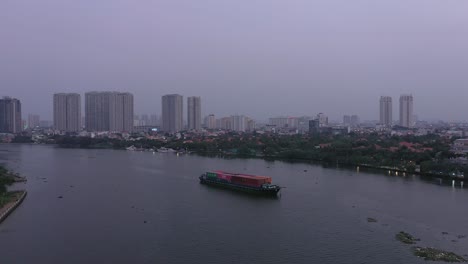 Container-boat-carrying-cargo-on-Saigon-River,-Vietnam-at-sunset-with-drone-reveal-of-city-skyline