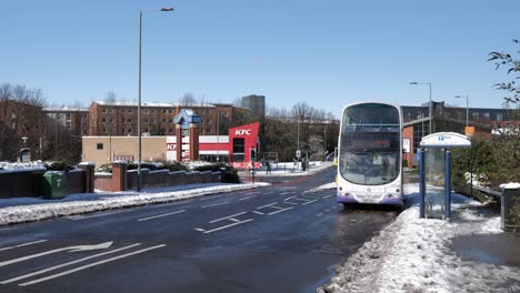 Autobús-Que-Sale-De-La-Parada-De-Autobús-Y-El-Tráfico-Pasa-Por-Una-Carretera-Soleada-Y-Nevada,-Sheffield