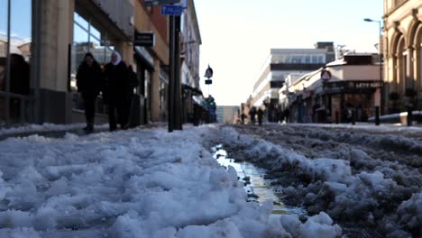People-walk-along-snowy,-slushy-pavement-toward-low-angle-camera-on-sunny-day