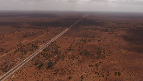 Cinematic-shot-of-white-van-on-tour-driving-on-empty-highway-surrounded-by-red-barren-nature-of-Australia---drone-tracking-shot