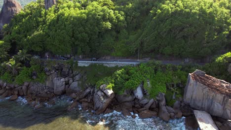 Low-drone-shot-of-passing-cars-on-road-near-cliff,-huge-granite-rock-at-Anse-forbans-beach-Mahe-Seychelles