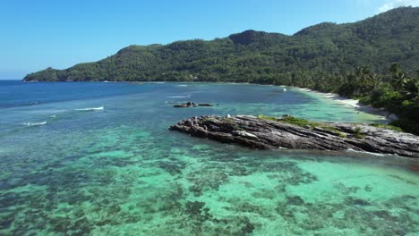 Low-flying-drone-over-large-granite-rock-at-Anse-forbans-beach,-locals-fishing,-people-swimming-at-sea-Mahe-Seychelles