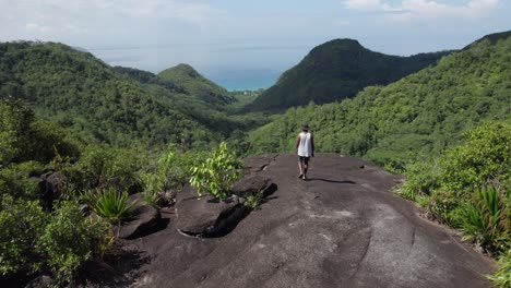 Drone-shot-of-man-walking-to-the-edge-of-the-cliff-inside-the-dense-tropical-forest-on-Mahe-island