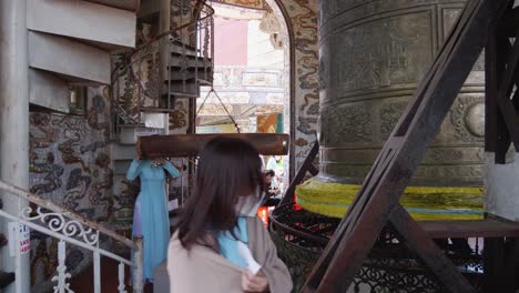 Mujer-Con-Vestido-Azul-Golpea-La-Campana-Budista-Con-Un-Tronco-Suspendido-Dentro-Del-Templo-Linh-Phuoc-En-Vietnam