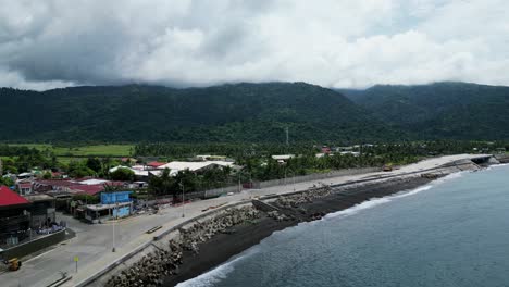 Aerial-View-of-idyllic-sea-waves-crashing-against-coastal-rural-town-with-newly-built-breakwater,-and-mountainous-background