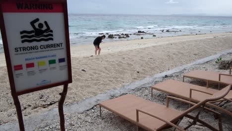 Nahaufnahme-Eines-Warnschilds-Mit-Starker-Strömung-Am-Strand-Und-Leerer-Sonnenliegen-Am-Strand-Mit-Einem-Einsamen-Touristen-Im-Hintergrund-In-Cebu,-Philippinen