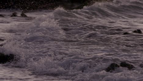 Closeup-shot-of-waves-rolling-up-on-beach-in-Hawaii,-splashing-over-rocks-at-sunset