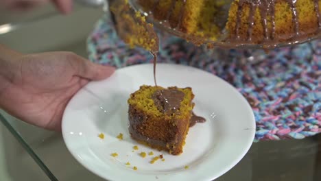 Serving-a-simple-homemade-carrot-cake-with-chocolate-sauce-on-a-white-plate-with-a-metal-spatula-lifting-the-piece-of-candy-with-the-syrup-dripping-onto-the-plate