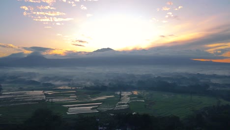 Aerial-view-of-orange-cloudy-sunrise-sky-and-mountain-with-slightly-fog-covered-the-plantation-and-rice-field