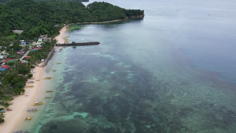 Steigende-Luftaufnahme-Des-Unberührten-Meereswassers-Mit-Blick-Auf-Bangka-Boote,-Die-An-Einem-Idyllischen-Weißen-Sandstrand-Aufgereiht-Sind,-Der-Mit-Einer-Ländlichen-Gemeinde-In-Catanduanes,-Philippinen,-Verbunden-Ist