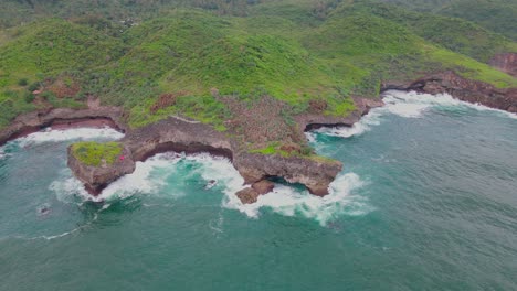 Waves-reaching-rocky-coastline-with-overgrown-island-in-Indonesia---Drone-top-down