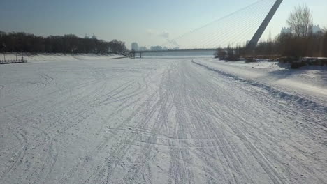 Low-aerial-flight-along-tracked-frozen-river-surface-to-cable-bridge