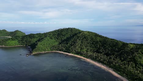 Stunning-Aerial-Panning-shot-of-jungle-covered,-mountainous-island-with-white-sand-beach-and-crystal-clear-sea-waters