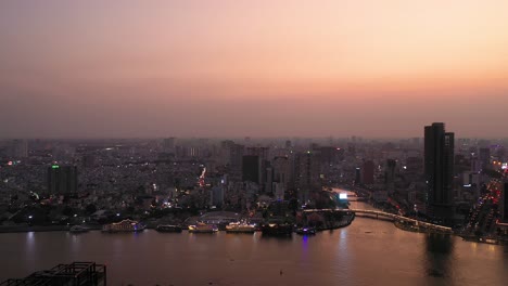 Ho-Chi-Minh-City,-Vietnam-iconic-Skyline-and-Saigon-river-waterfront-aerial-panning-shot-on-a-busy-evening-featuring-key-buildings-illuminated-against-beautiful-colored-sky-and-reflected-in-the-river