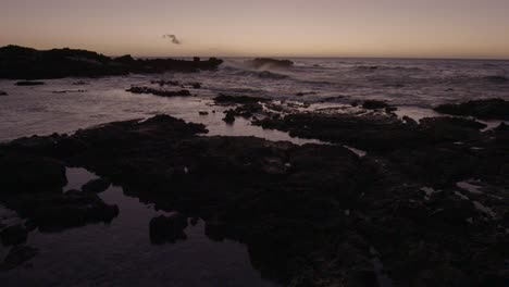 Static-shot-of-Hawaii-beachfront-at-dusk,-as-waves-crash-into-rocks-in-the-distant-ocean