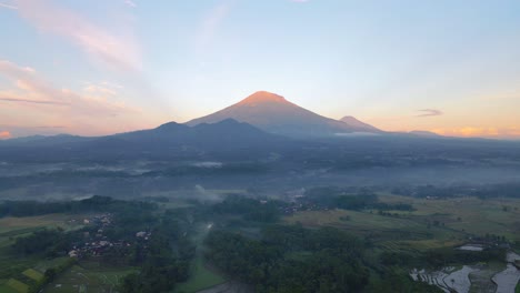 Aerial-footage-of-beautiful-rural-landscape-in-the-foggy-morning-with-view-of-countryside,-plantation-and-huge-mountain-and-orange-sky