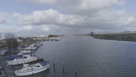 Drohnenaufnahme-Eines-Kleinen-Hafens-An-Einem-Fluss-Mit-Windmühle-Im-Hintergrund