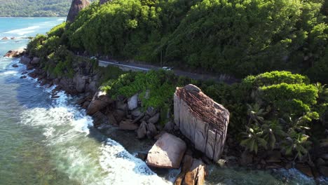 Side-drone-shot-of-passing-cars-on-road-near-cliff,-huge-granite-rock-at-Anse-forbans-beach-Mahe-Seychelles