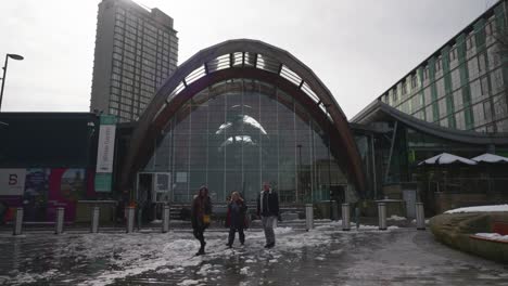 People-walking-in-the-street-past-the-Winter-Garden-attraction-in-Sheffield-city-centre-during-snowy,-winter-time