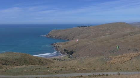Paragliders-soar-above-a-barren-landscape-in-contrast-to-beautiful-blue-ocean-in-summertime---Godley-Head,-Banks-Peninsula