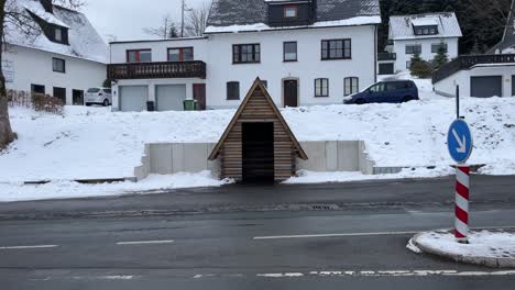 Kleines-Holzhaus-Auf-Einer-Straße-Mit-Vorbeifahrenden-Autos-Im-Schnee-Im-Winter-Mit-Wohnhäusern-Im-Hintergrund