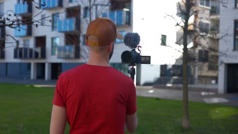A-Caucasian-man-vlogs-with-a-DSLR-and-external-mic,-capturing-a-selfie-style-video-in-front-of-apartments