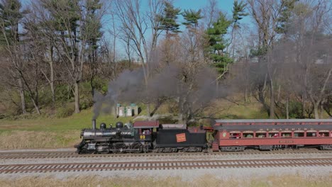 A-Drone-Side-View-of-a-Antique-Steam-Passenger-Train-Blowing-Smoke,-Traveling-Thru-the-Farmlands-on-a-Sunny-Fall-Day