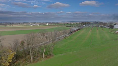 Drone-View-of-a-Steam-Passenger-Train-Approaching-Blowing-Smoke,-Traveling-Thru-the-Farmlands-on-a-Sunny-Fall-Day