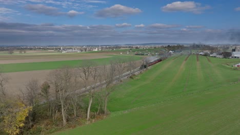 Vista-De-Drones-De-Un-Tren-De-Pasajeros-De-Vapor-Acercándose-A-Soplar-Humo,-Viajando-A-Través-De-Las-Tierras-De-Cultivo-En-Un-Día-Soleado-De-Otoño