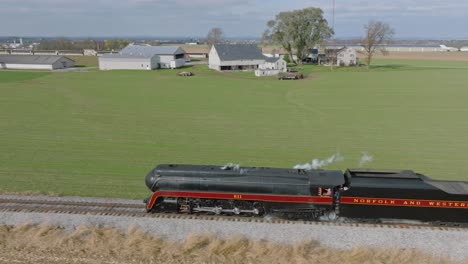 A-Drone-Side-View-of-a-Steam-Passenger-Train-Blowing-Smoke,-Traveling-Thru-the-Farmlands-on-a-Sunny-Fall-Day