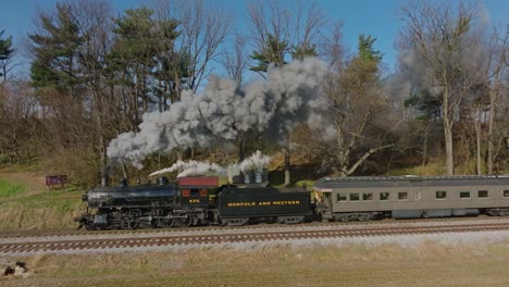 A-Drone-Above-and-to-the-Side-View-of-A-Restored-Steam-Passenger-Train-Passing-a-Small-Station-Blowing-Smoke-on-a-Sunny-Winter-Day